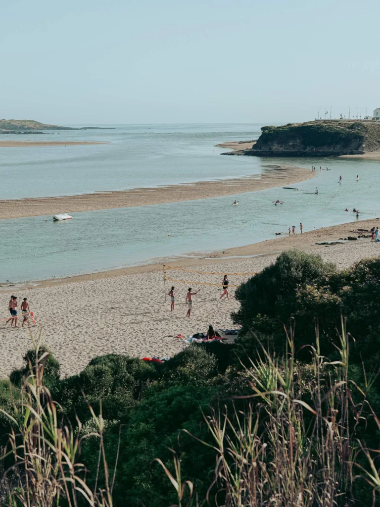 Vila Nova de Milfontes beach in Alentejo, Portugal, with people playing volleyball and swimming near the estuary under a clear sky in June.