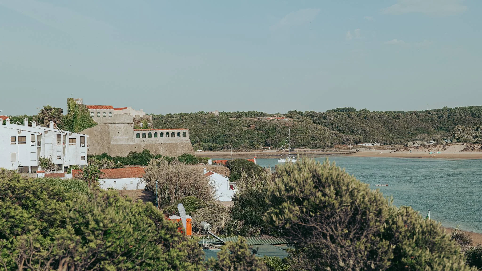 View of the Vila Nova de Milfontes fort and the Mira River, with white houses and a beach in the background under a clear sky.