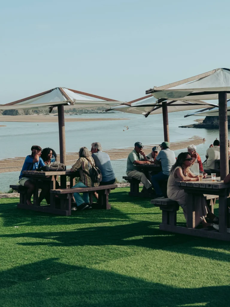 Groups of people enjoy an outdoor aperitif at wooden tables, under umbrellas, with a view of a river and the beach in Vila Nova de Milfontes, in Alentejo, Portugal.