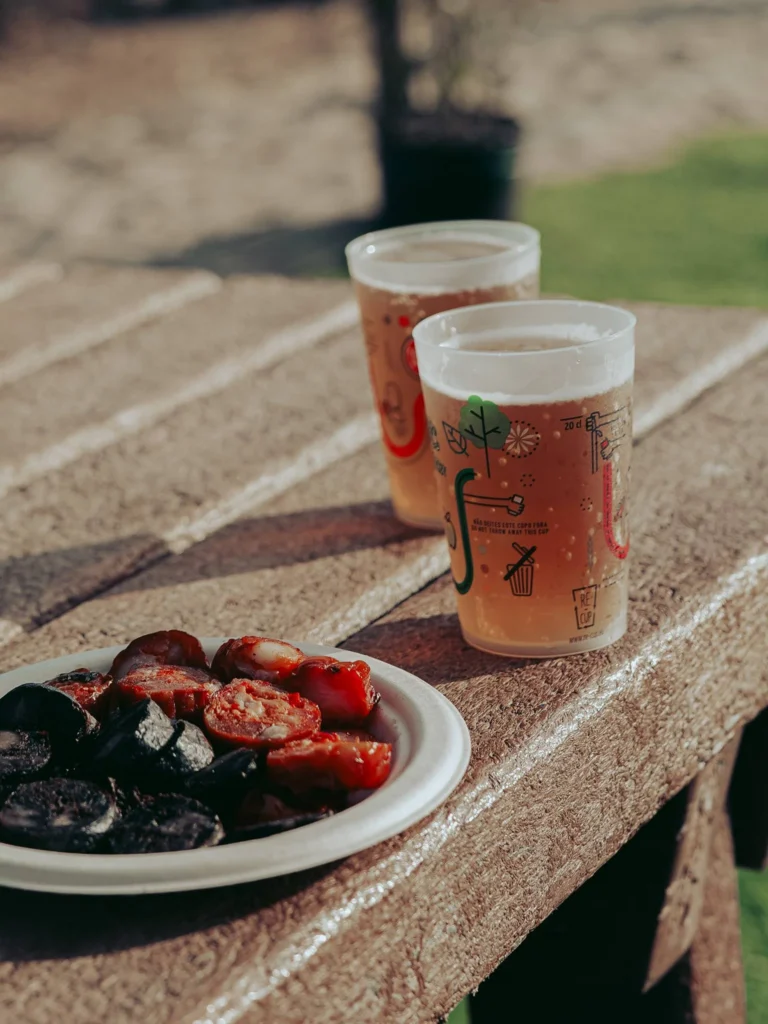Two glasses of beer and a plate of black and red chorizo are placed on an outdoor table during an aperitif in Vila Nova de Milfontes during the Fai-Tur festival, under a summery natural light.