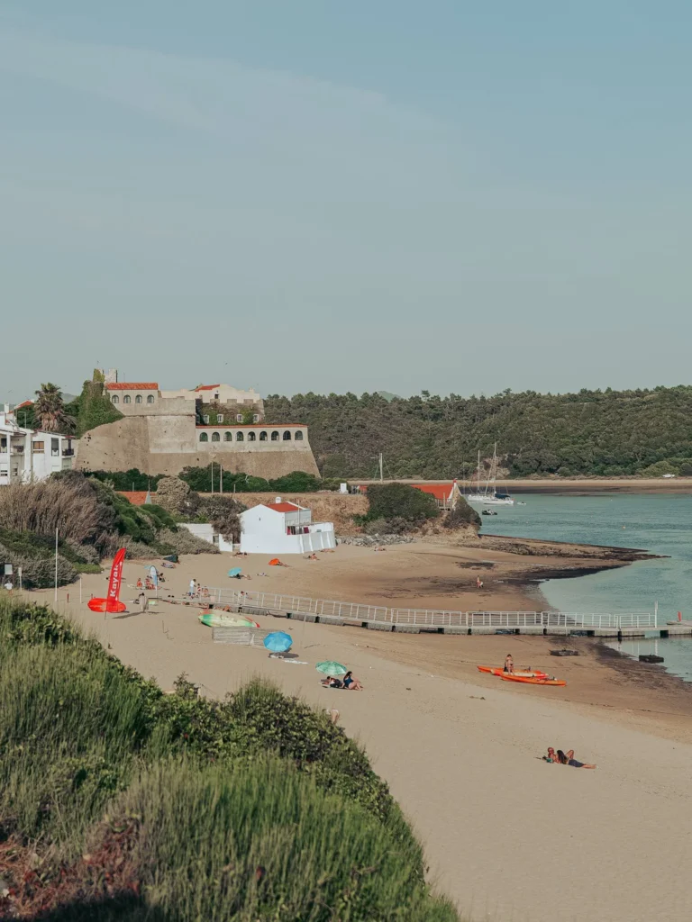 Sandy beach in Vila Nova de Milfontes with some colorful umbrellas, overlooking a fortified building and vegetation in the background, under a clear sky.