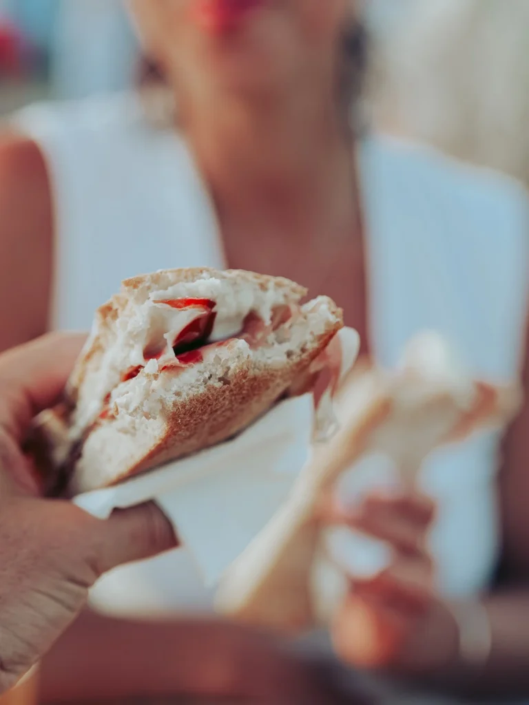 Close-up of a presunto sandwich with buttery cheese held in hand, with a slightly blurred woman in the background at a stand in Vila Nova de Milfontes in June.