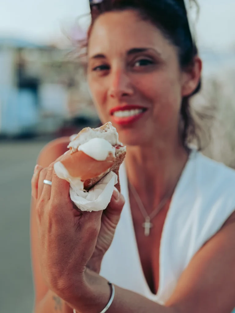 Caroline is holding a presunto and cheese sandwich in front of an outdoor stall in Vila Nova de Milfontes during a summer festival.