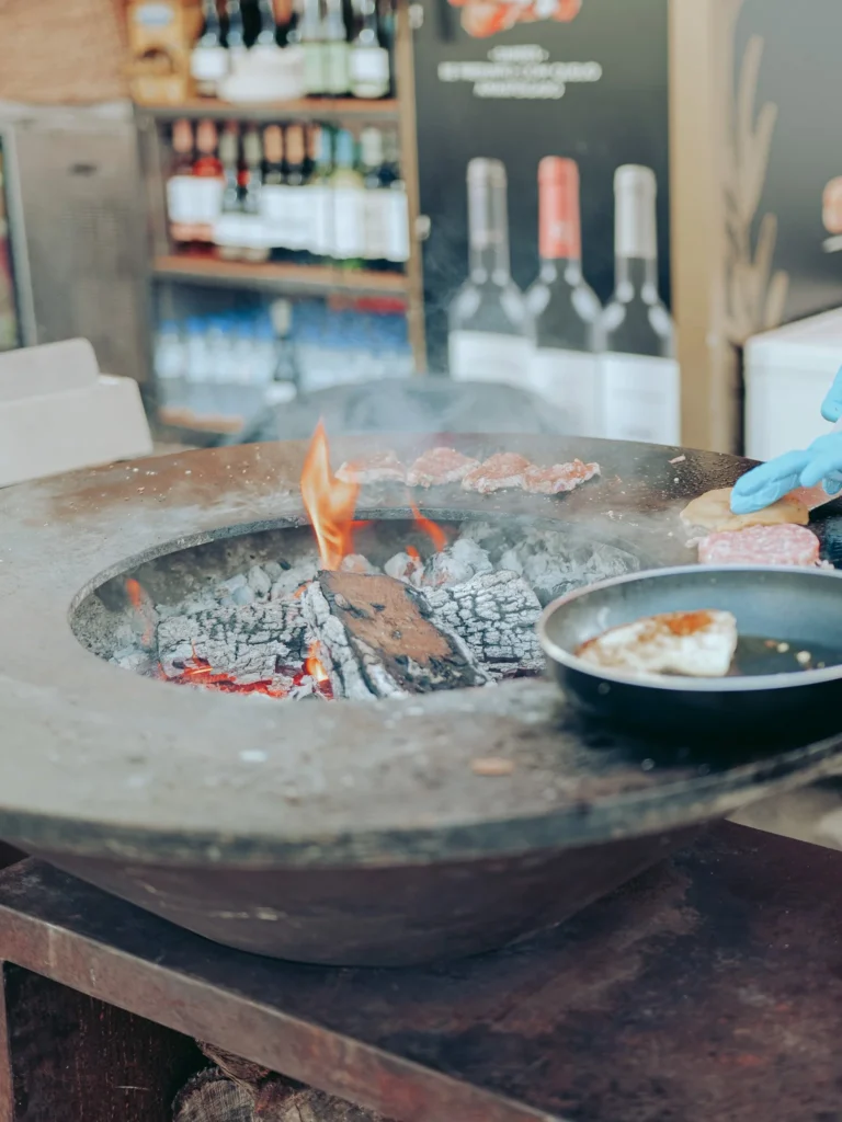 Close-up of an outdoor food stall in Vila Nova de Milfontes, with a wood fire grilling meat and cheese, and bottles of wine visible in the background.