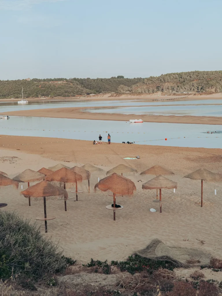 Vila Nova de Milfontes beach with straw umbrellas, calm lagoon and green hills in the background, under a clear sky in June.