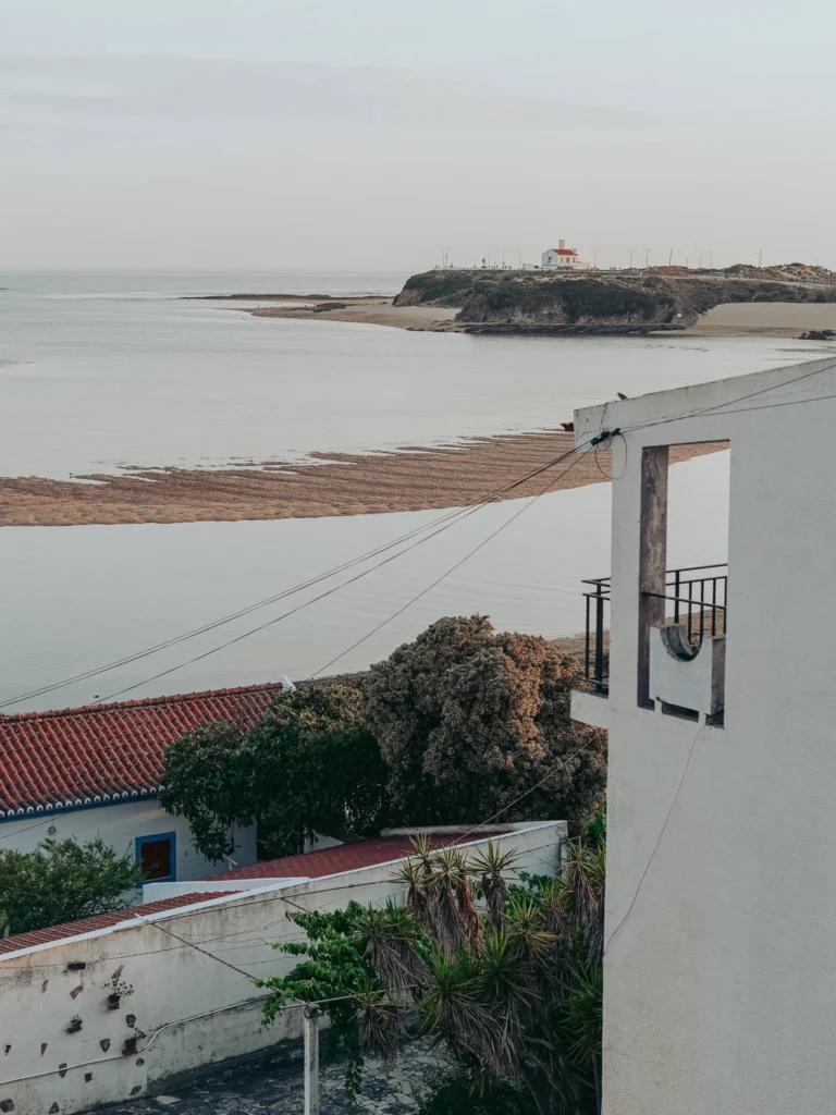 View of the estuary and coast of Vila Nova de Milfontes, with a white building in the foreground and a lighthouse in the distance on a rocky point. The atmosphere is calm at the end of the day.