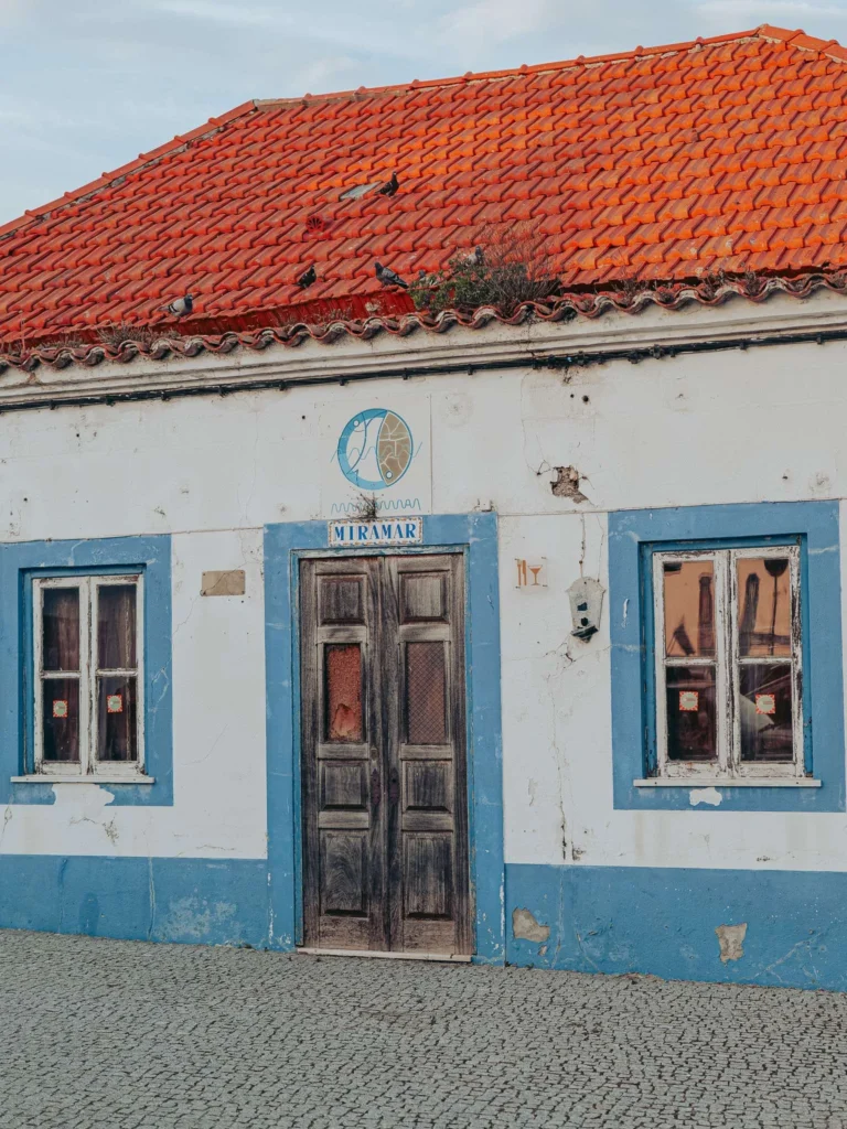 Facade of a traditional house in Vila Nova de Milfontes, with a red tiled roof, blue window frames, and a Miramar sign above the door.
