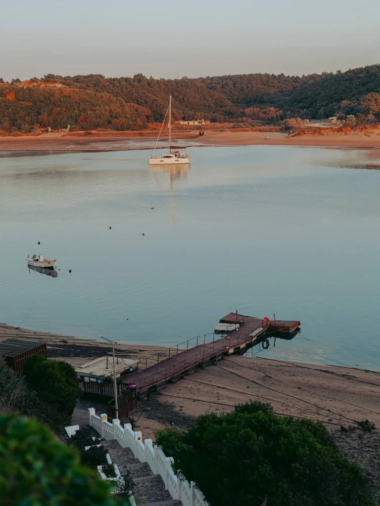 A wooden dock descends towards a peaceful beach, with a sailboat and a small boat moored on a river surrounded by lush hills in Vila Nova de Milfontes, Alentejo, Portugal, at the end of the day.