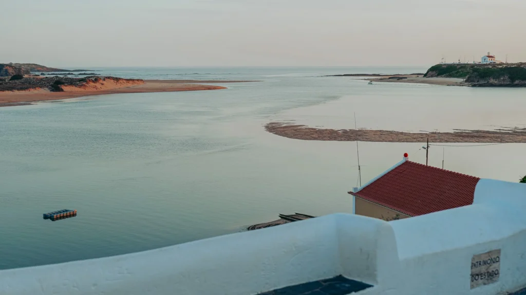 View of the estuary of Vila Nova de Milfontes with a sandy beach, a building with a red roof, and the sea in the background, at the end of a summer day.