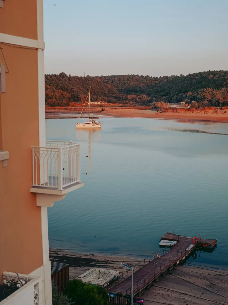 View of a peaceful river in Vila Nova de Milfontes with a moored catamaran and a wooden jetty in the foreground, at the end of a summer day.