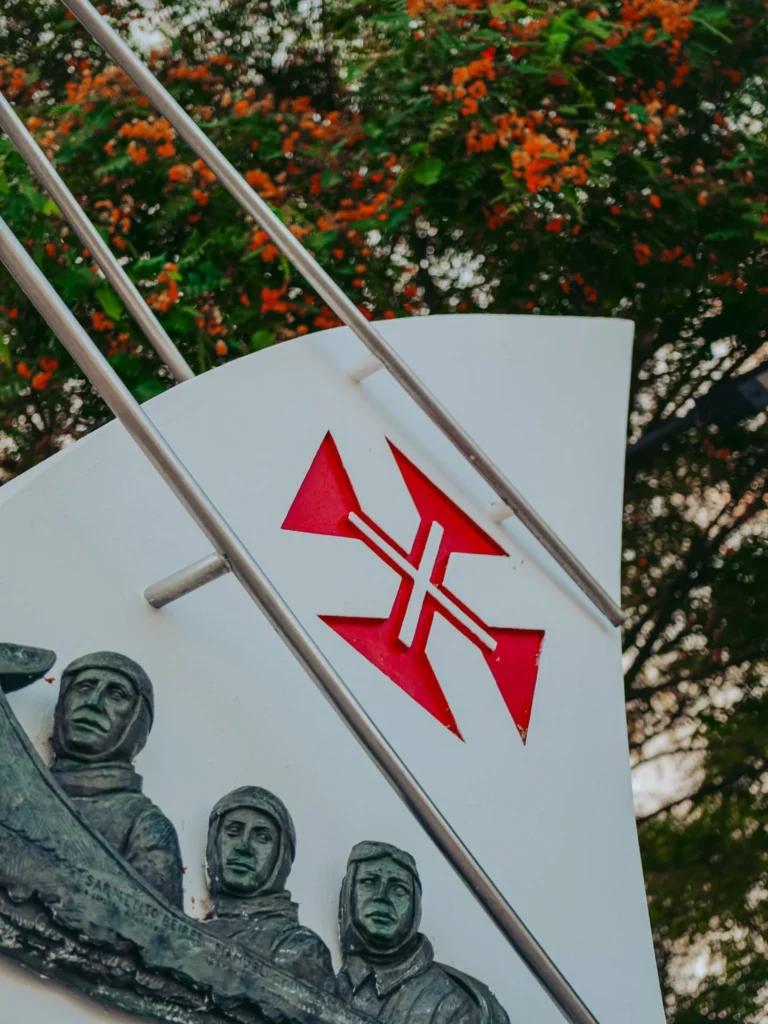 Detail of the monument to aviators Gago Coutinho and Sacadura Cabral in Vila Nova de Milfontes: red cross of the Order of Christ and sculpted faces recalling the first aerial crossing of the South Atlantic in 1922, with flowering trees in the background.