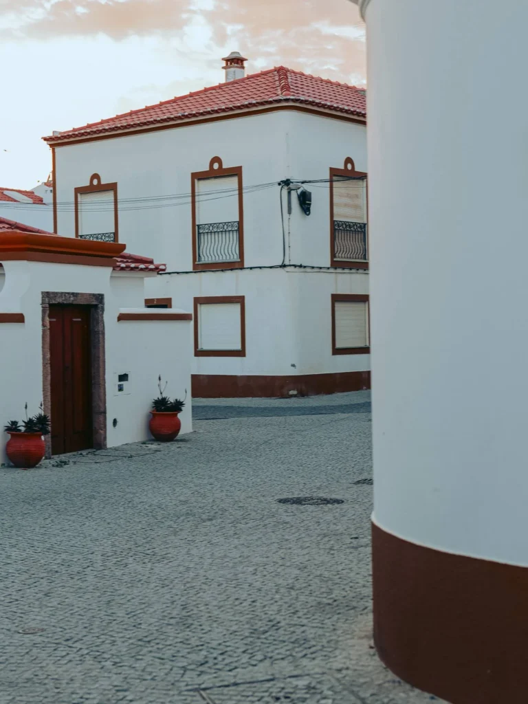 Cobblestone alley lined with white houses with red roofs typical of Alentejo in Vila Nova de Milfontes, under a slightly cloudy sky.