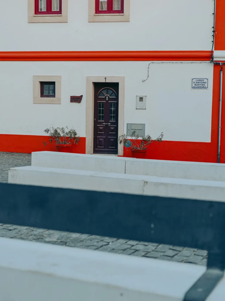 Facade of a white and red house in Vila Nova de Milfontes, with a dark door, framed windows, and potted plants in front of the entrance.