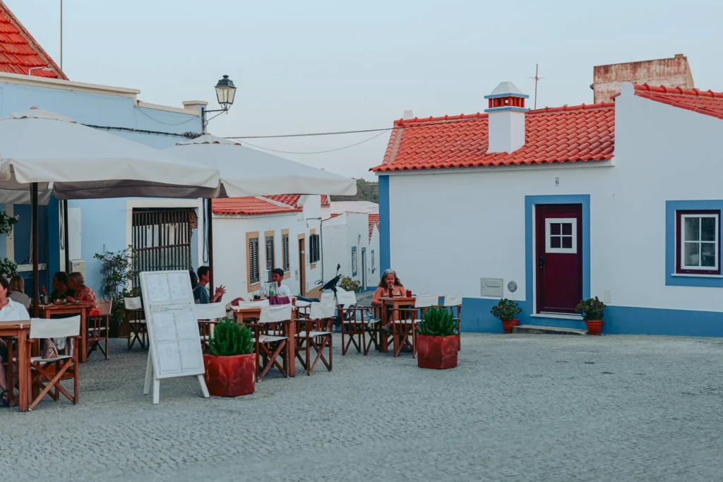 Outdoor café terrace on a cobbled square, surrounded by traditional houses with red roofs in Vila Nova de Milfontes, Alentejo, on a June day.