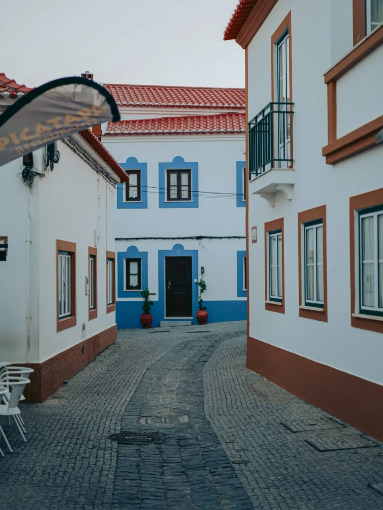 Cobblestone alley lined with white houses featuring colorful frames typical of Vila Nova de Milfontes, in Alentejo, Portugal, photographed in June.