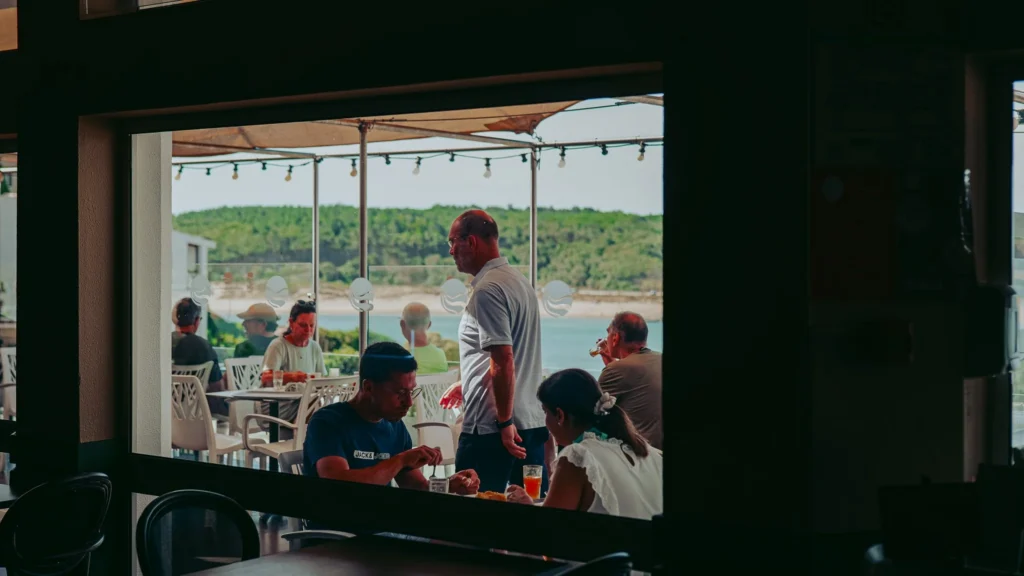 Restaurant terrace with several people seated for breakfast, overlooking the river and greenery in the background in Vila Nova de Milfontes.