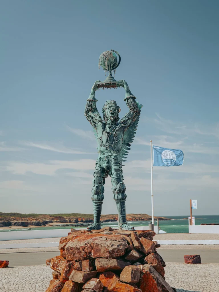 Sculpture "Arcanjo" from Vila Nova de Milfontes: a figure made of recycled iron holding a globe above its head, standing on a stone pedestal near the Miradouro do Farol facing the Atlantic Ocean, under a clear sky in Alentejo.