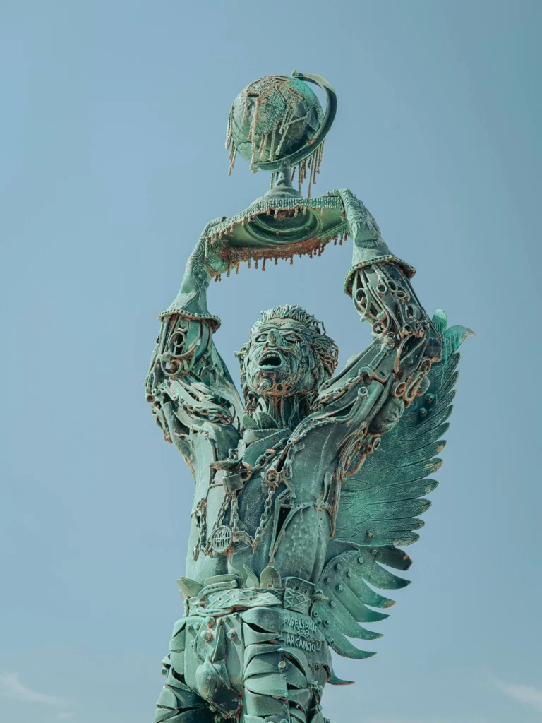 Sculpture "Arcanjo" from Vila Nova de Milfontes: winged figure made of recycled iron holding a globe above its head, an urban artwork with an ecological message by artist Aureliano de Aguiar, photographed under a clear sky in Alentejo.