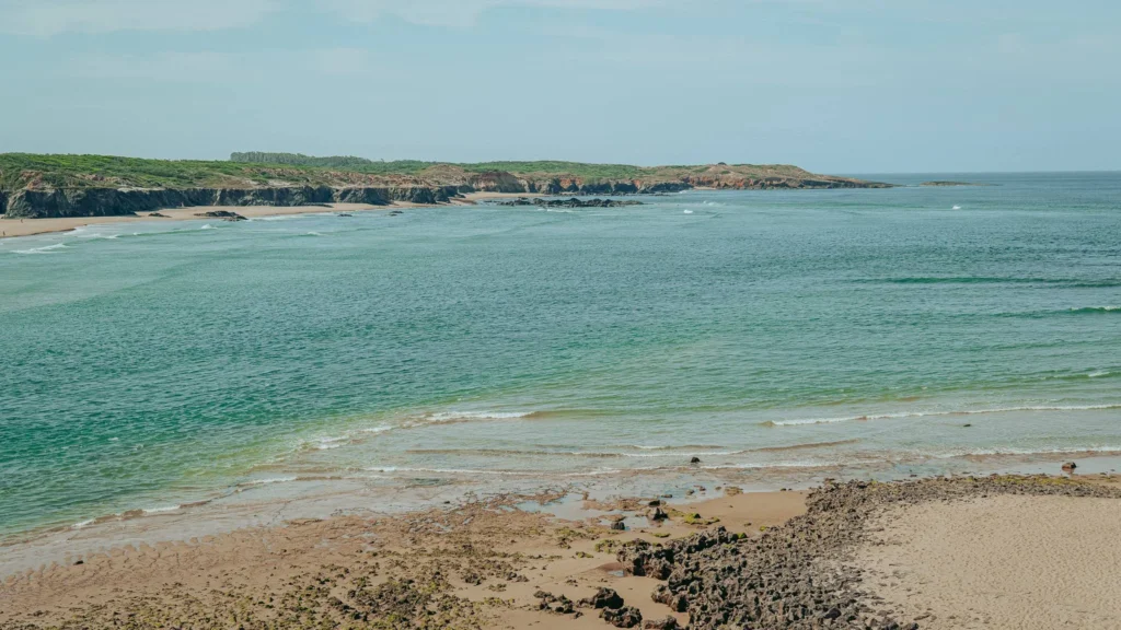 Wild beach with sand, rocks, and turquoise water on the coast of Vila Nova de Milfontes, in Alentejo, Portugal, under a clear sky in June.