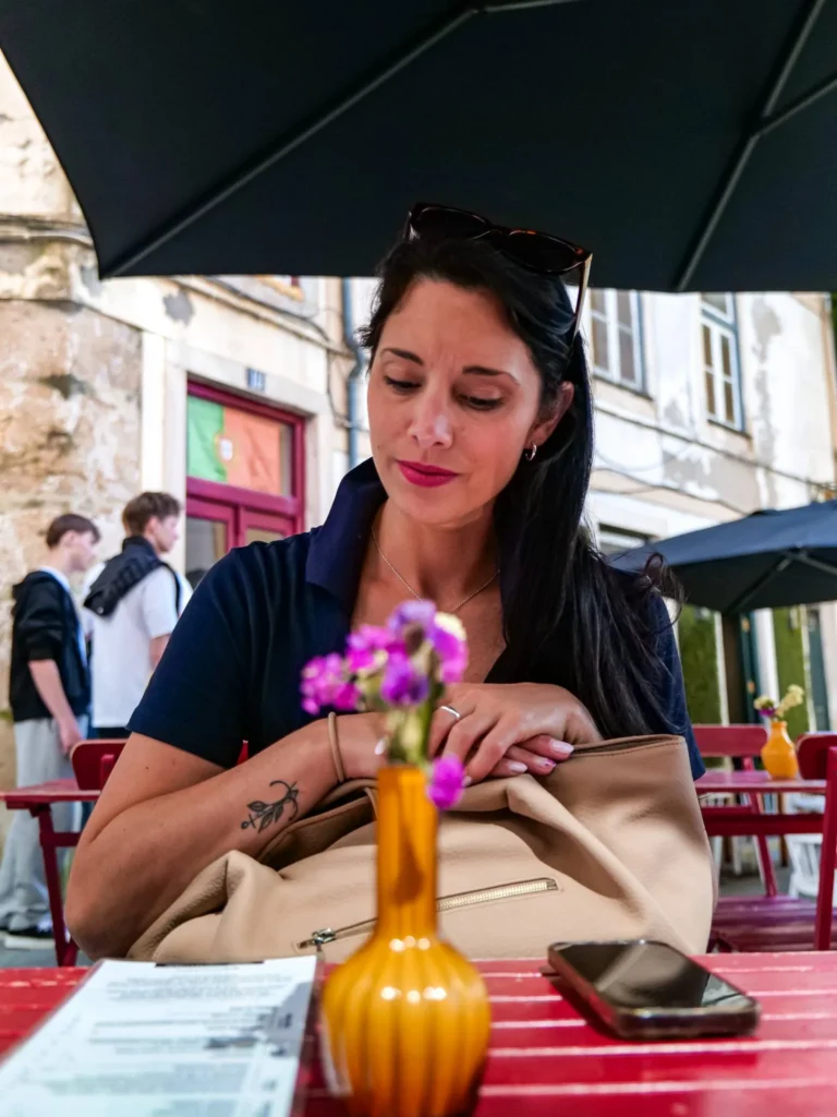 Caroline sitting at a café terrace in Coimbra, Portugal, under a large black umbrella, with a menu and a vase of flowers on the table.