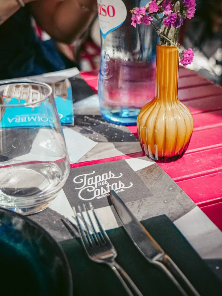 Table set on the terrace in Coimbra with a carafe of water, a glass, cutlery, and a menu from the restaurant Tapas nas Costas, accompanied by a small vase of flowers.