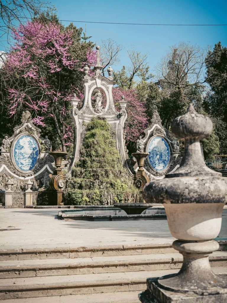 Baroque fountain adorned with azulejos and sculptures in the Sereia garden in Coimbra, surrounded by flowering trees in spring.