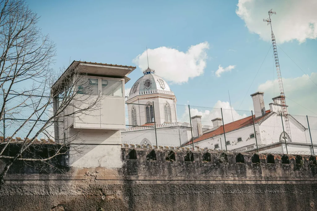 Exterior view of a historic prison in Coimbra, Portugal, featuring a watchtower, barbed wire, and old buildings under a clear sky.