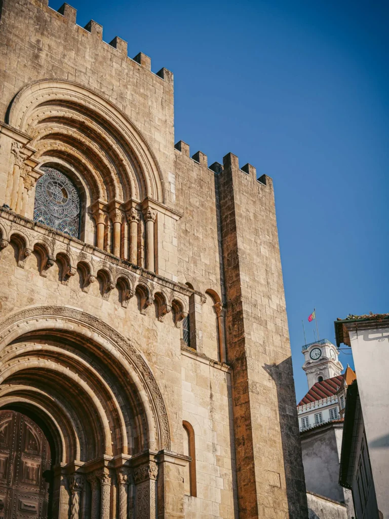 Facade of the Sé Velha in Coimbra under a blue sky, with the university tower and the Portuguese flag visible in the background.