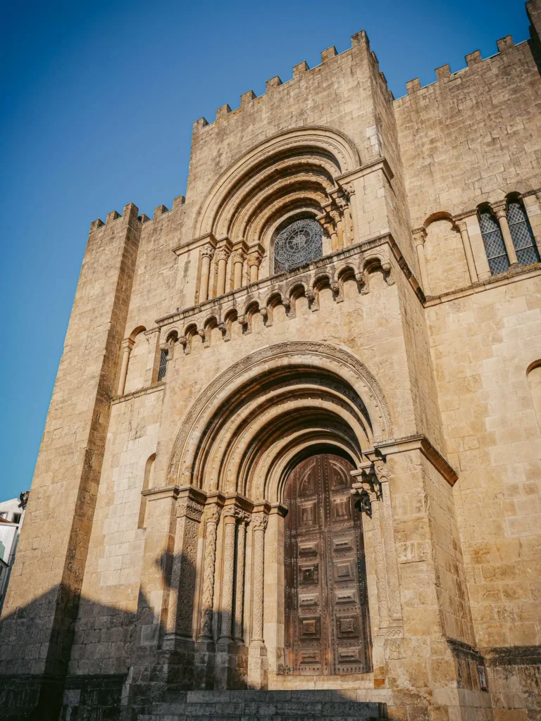 Facade of the Sé Velha de Coimbra, an imposing Romanesque stone cathedral under a blue sky, with no visible human presence.