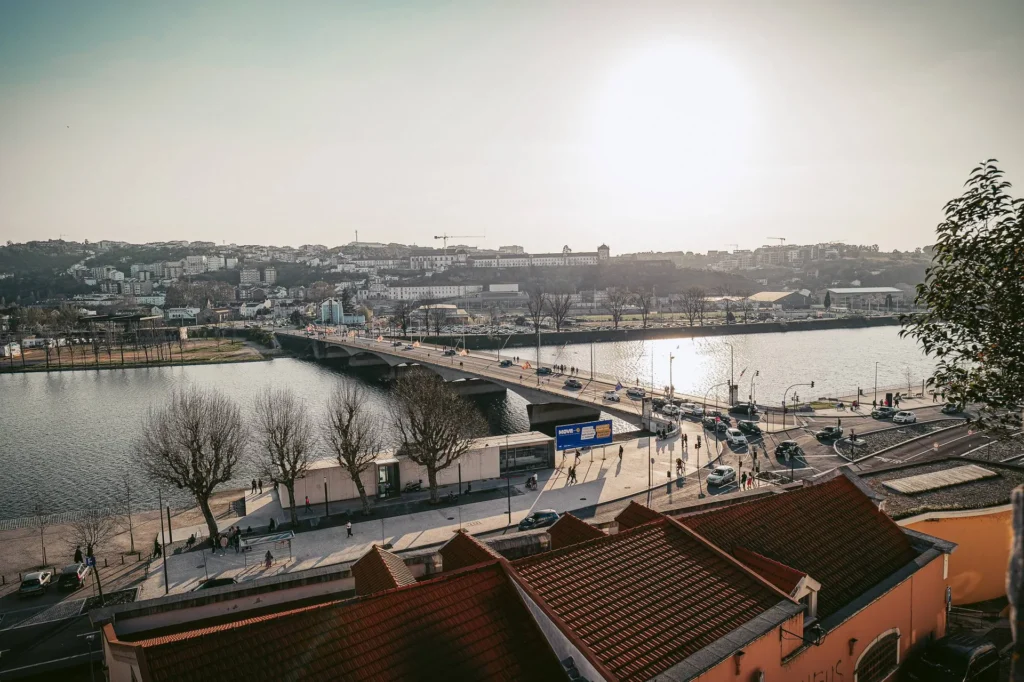 View of the main bridge of Coimbra spanning the Mondego River at sunset, with the city in the background and a few pedestrians on the quays.