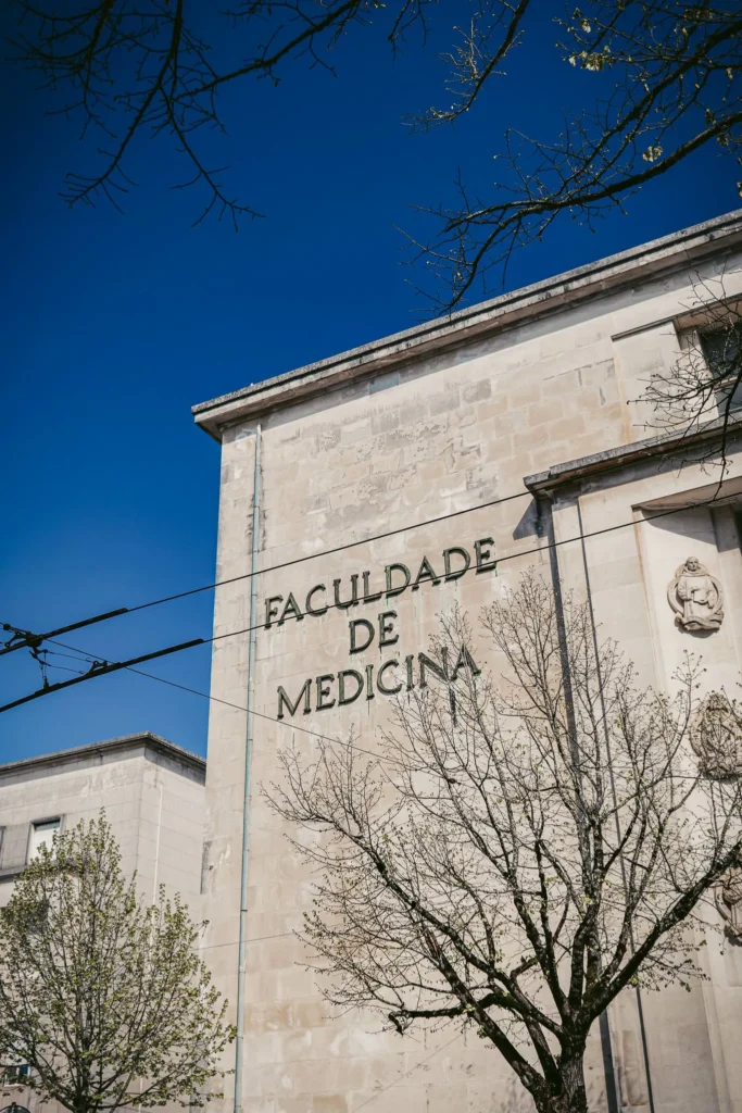 Facade of the Faculty of Medicine of Coimbra under a blue sky, with trees beginning to bloom.