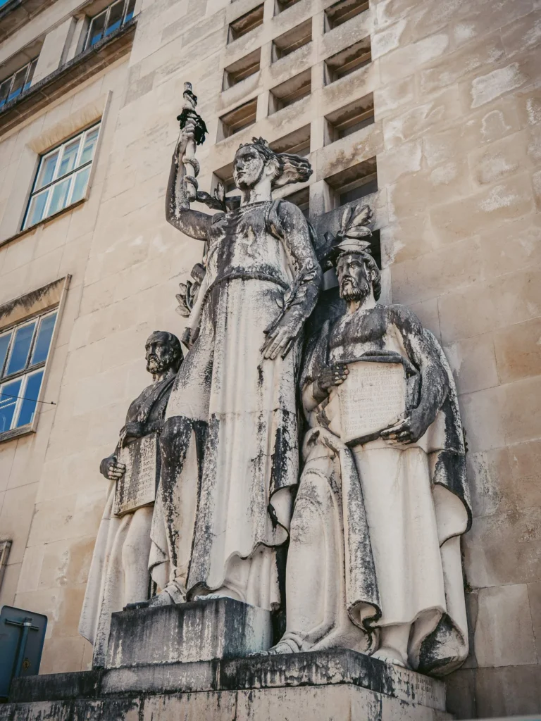 Monumental stone statues in front of a building facade in Coimbra, representing three allegorical figures, likely related to history or knowledge.