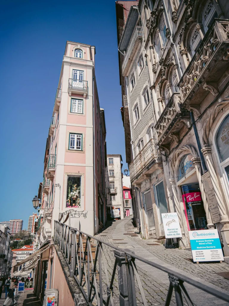 Narrow and steep street in the historic center of Coimbra, Portugal, lined with old buildings and local shops under a blue sky.