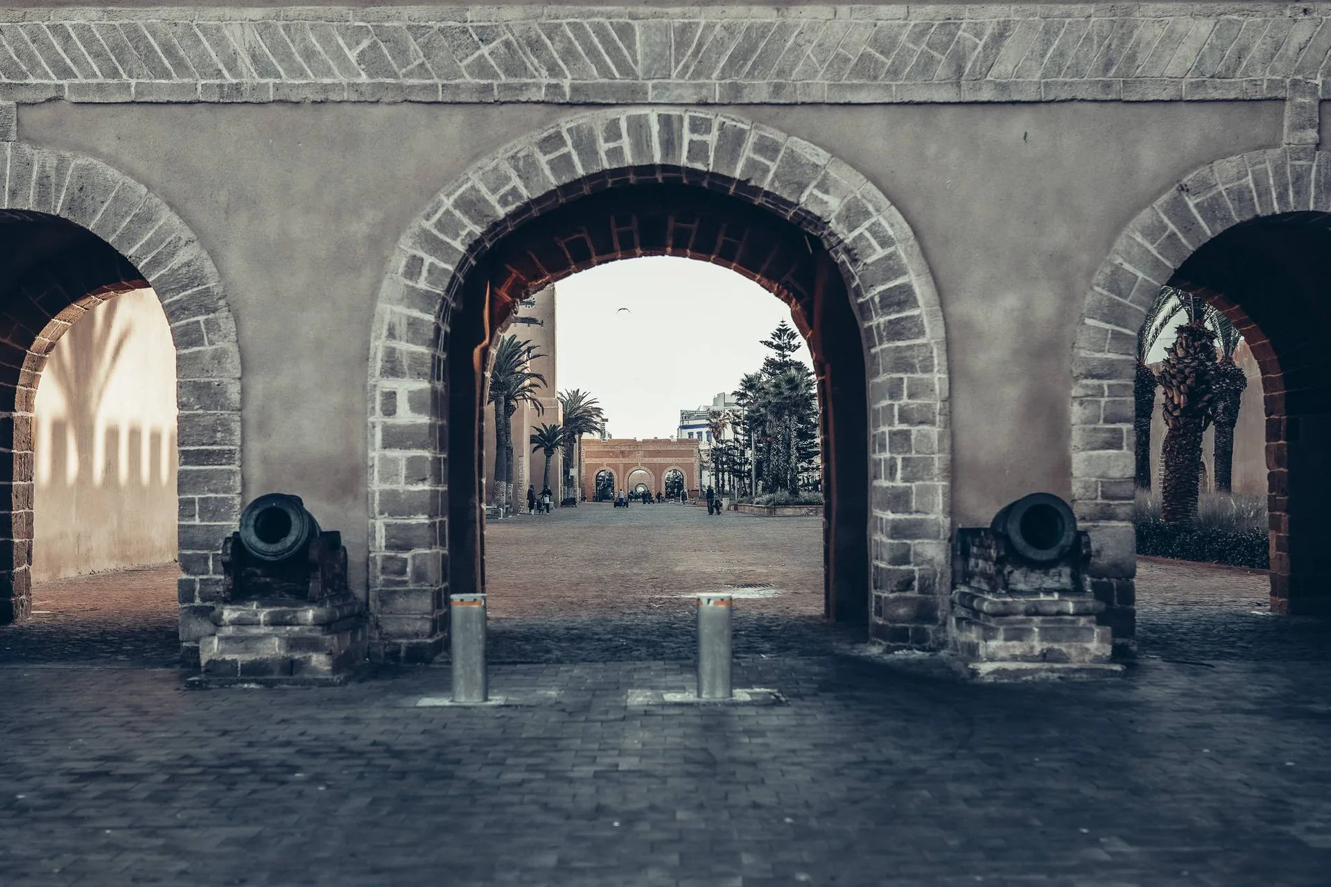 View through a fortified stone door in Essaouira, with ancient cannons and a palm-lined pathway in the background.