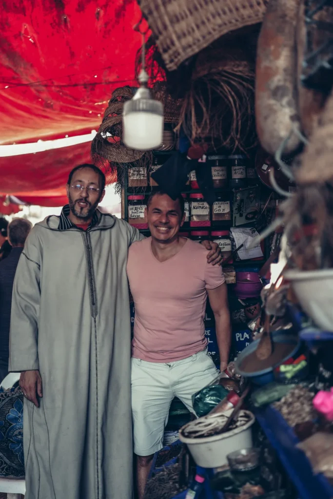 Xavier poses with Abdulhadi, an herbalist in a traditional shop in Essaouira, surrounded by jars of herbs and handcrafted items under a red tent.