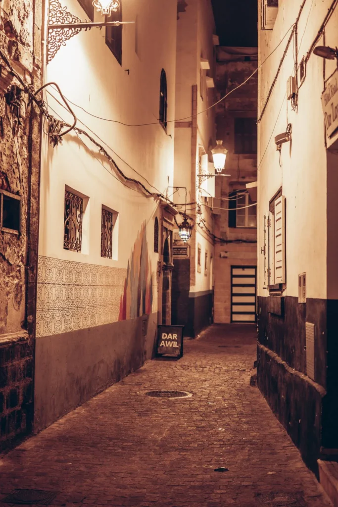 Narrow alley of the medina of Essaouira in Morocco, illuminated by street lamps in the evening, with white walls and traditional patterns.