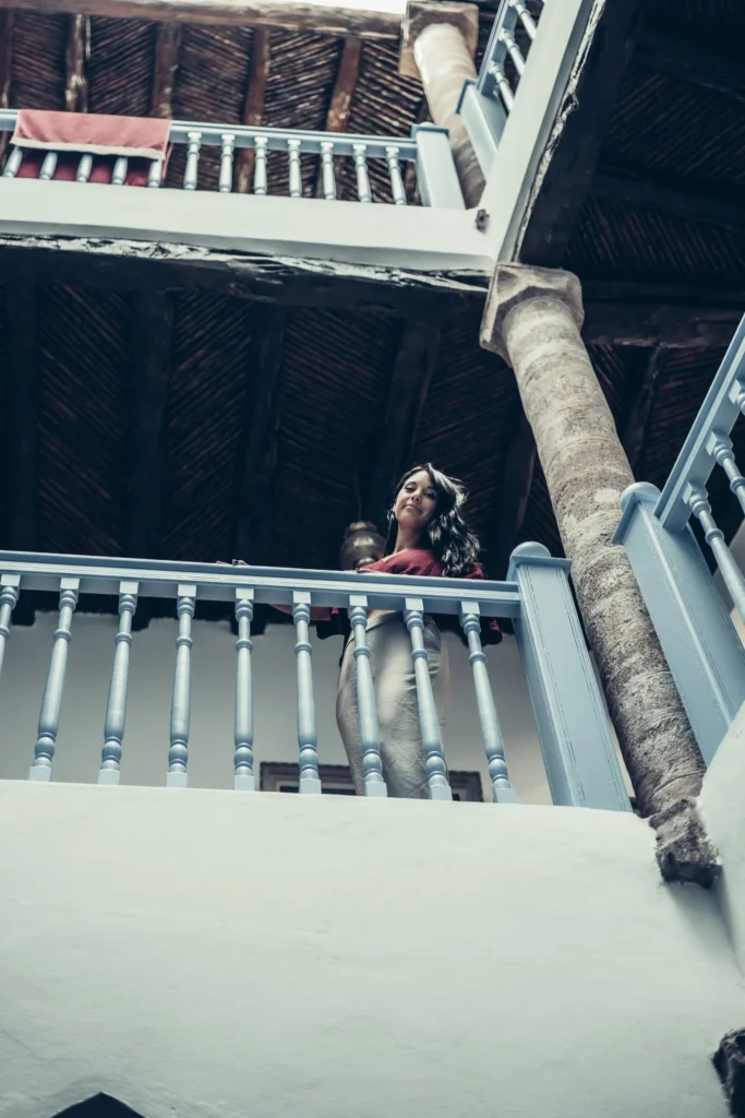 Caroline on the interior gallery of a traditional riad in Essaouira, with painted balustrades and stone columns under a wooden roof.