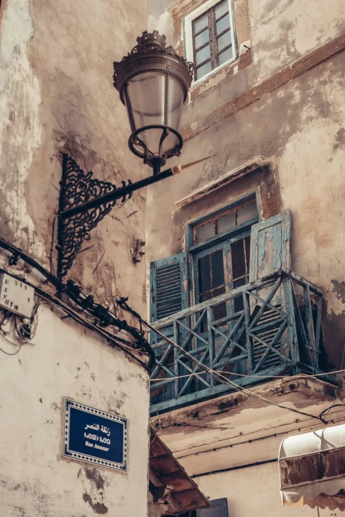 Facade of an old alley in Essaouira with a worn blue wooden balcony, a traditional street lamp, and a street sign 'Rue Ammar'.
