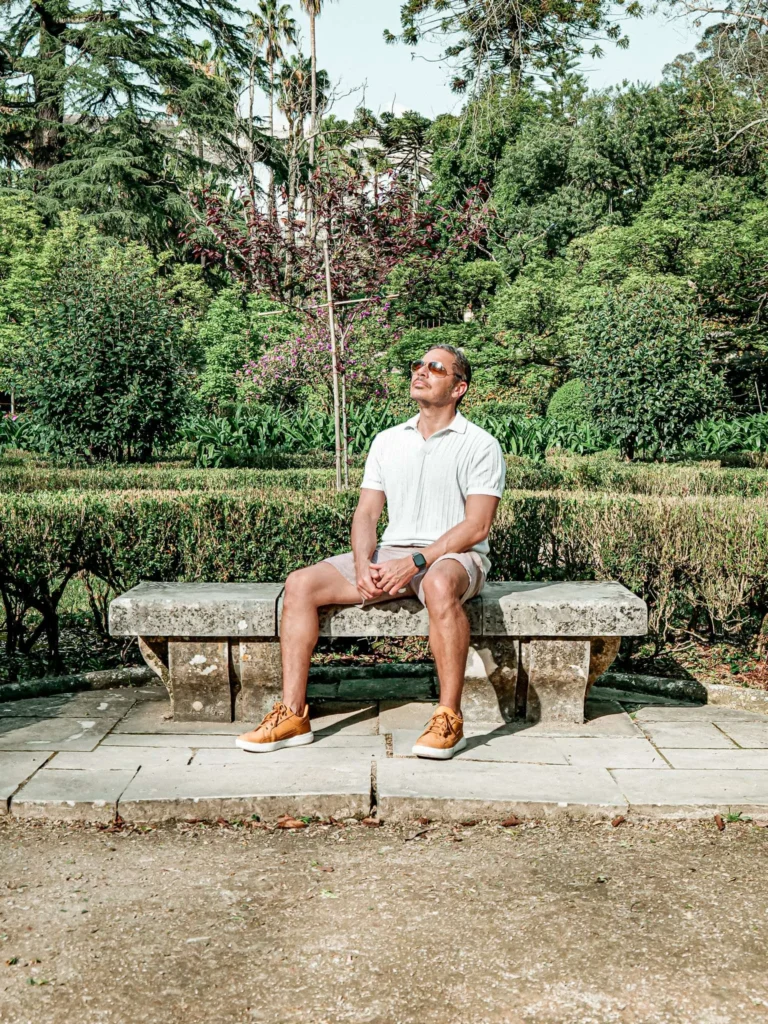 Xavier is sitting on a stone bench in the Botanical Garden of Coimbra, surrounded by lush vegetation at the beginning of spring.