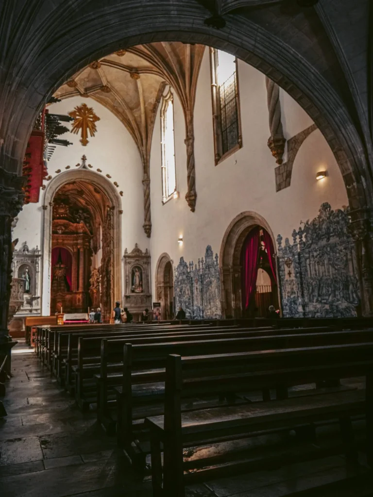 Interior of the Santa Cruz church in Coimbra, with wooden benches, azulejos on the walls, and a richly decorated altar. Natural light enters through large Gothic windows.