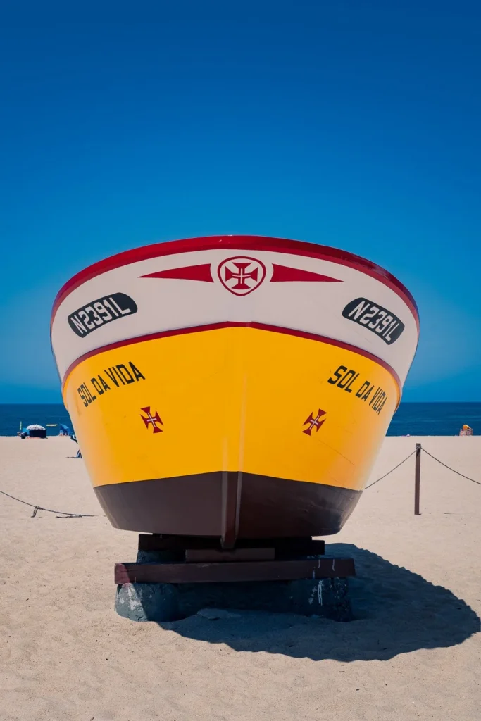 Traditional yellow and red boat resting on the sand of a beach under a blue sky in Nazaré, Portugal, in summer 2023.