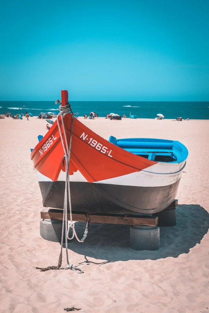 Traditional red and black boat resting on the sand of a Nazaré beach, with the Atlantic Ocean in the background and holidaymakers under umbrellas.
