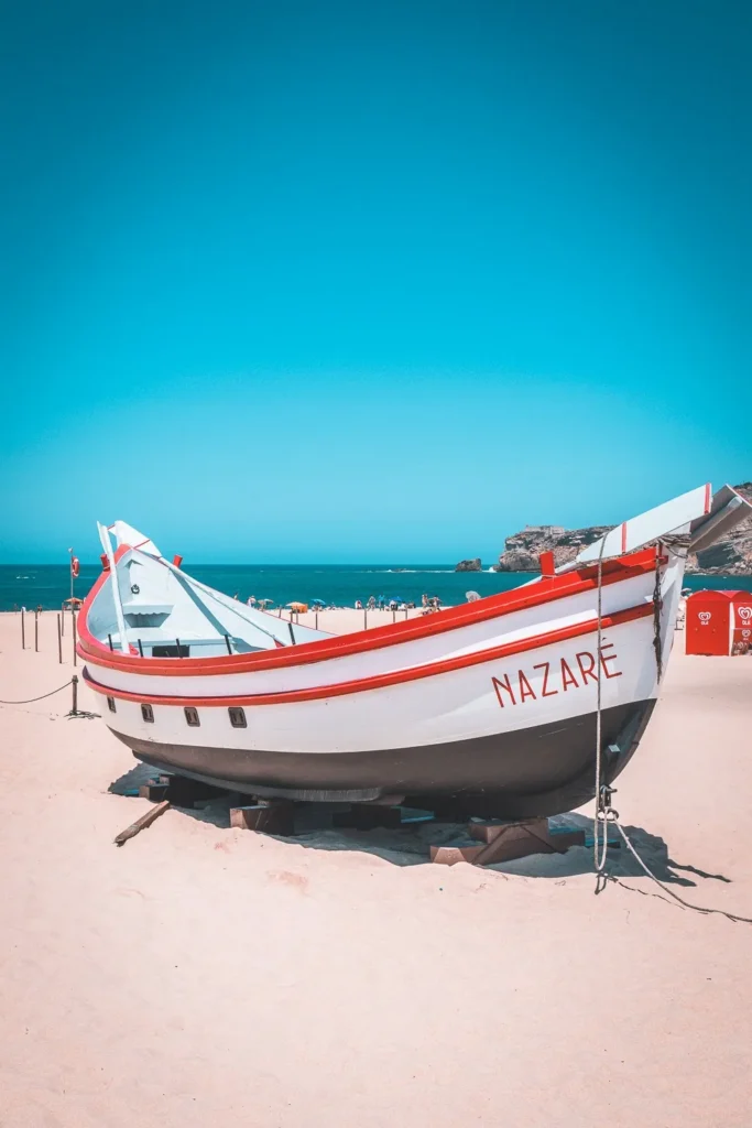Traditional fishing boat displayed on Nazaré beach under a blue sky, with the Atlantic Ocean in the background.