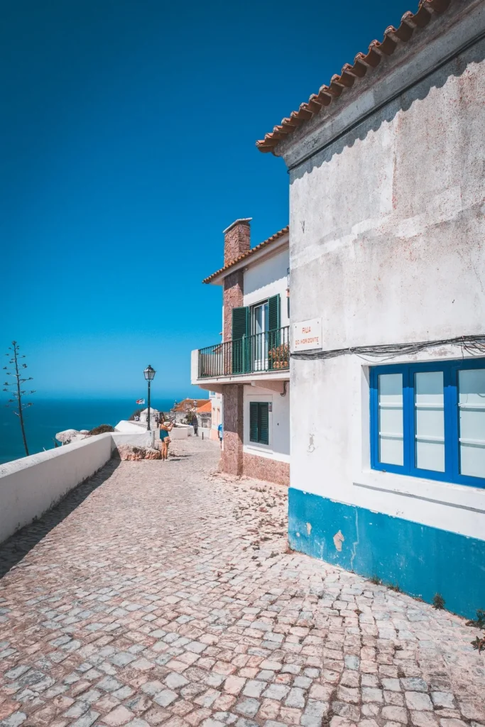 Cobblestone alley in Nazaré lined with white houses with colorful shutters, overlooking the Atlantic Ocean under a blue sky in July 2023.