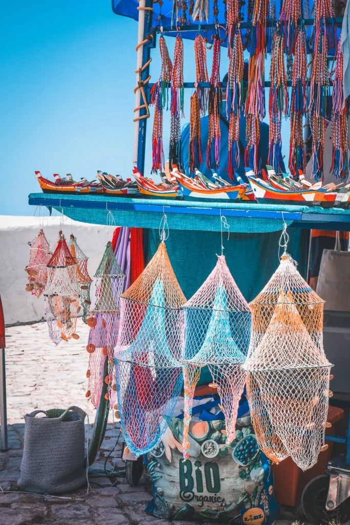 Market stall in Nazaré displaying colorful nets, traditional boat models, and handcrafted souvenirs under a blue sky in summer.