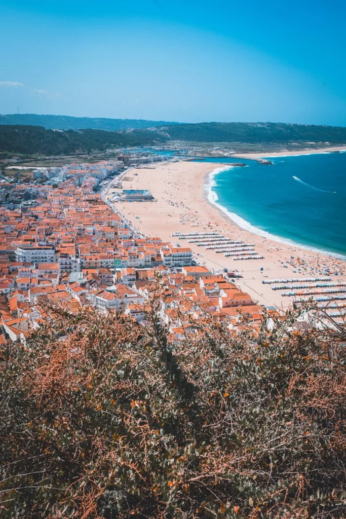 Panoramic view of Nazaré beach and the red rooftops of the village, with the Atlantic Ocean in the background under a clear summer sky.