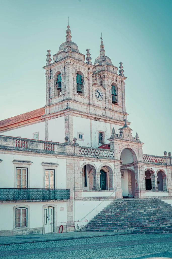 Facade of the baroque-style church of the Sanctuary of Our Lady of Nazaré, with its two towers and a stone staircase, under a clear summer sky in Nazaré.