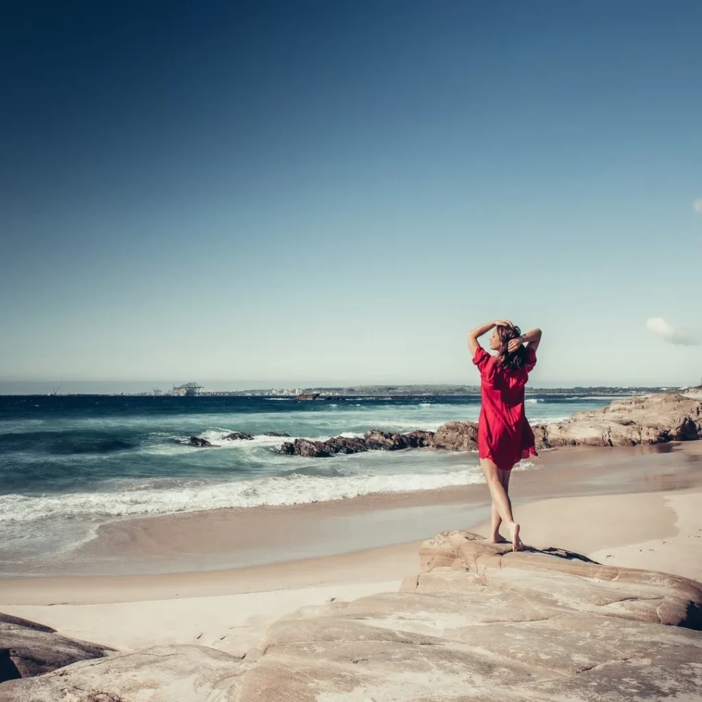 Caroline in a red dress walks on the rocks of Oliveirinha beach near Porto Covo, with the Atlantic Ocean in the background under a clear sky in March 2022.