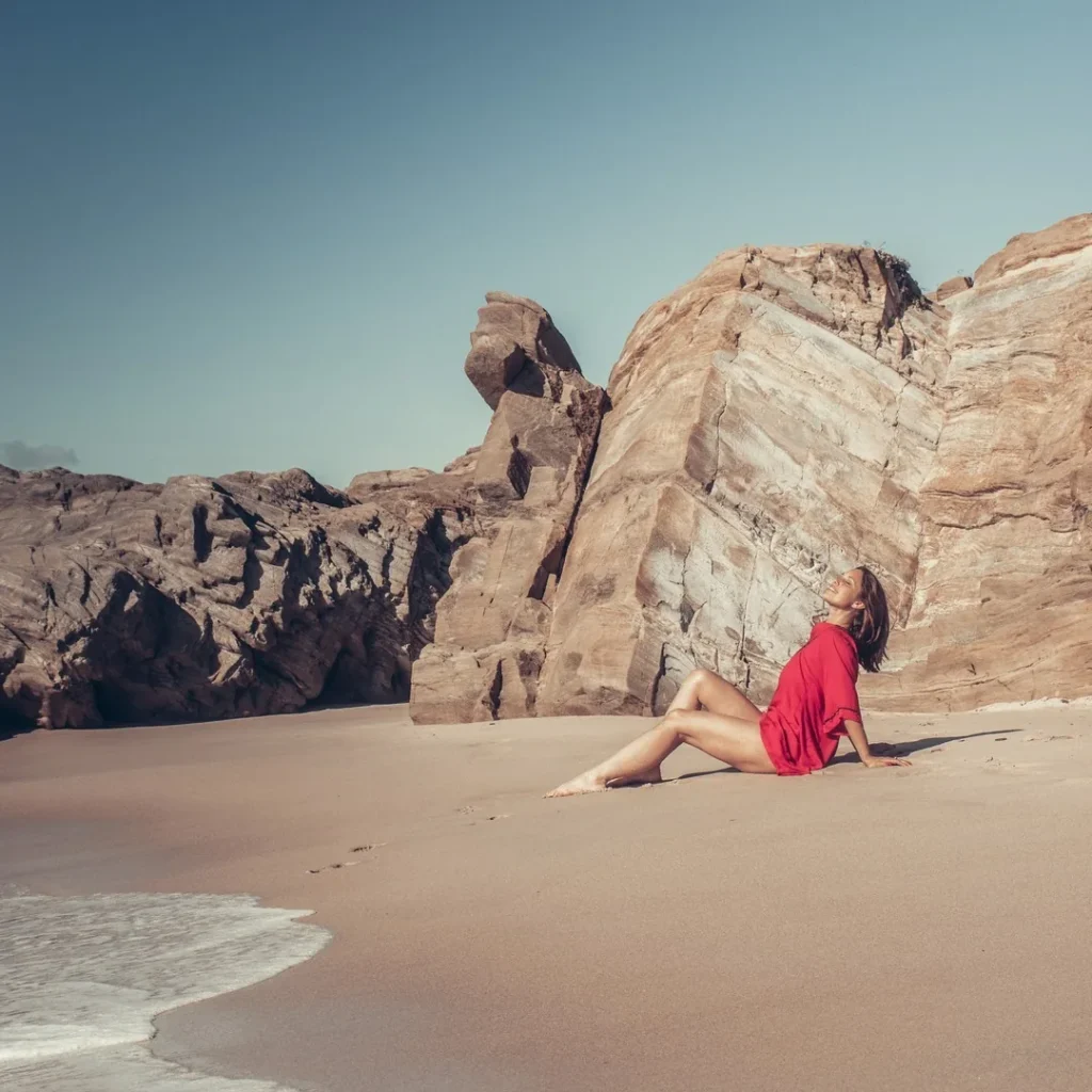 Caroline sitting on the sand in front of large rock formations at Praia de Oliveirinha, under the March sun of 2022 in the Alentejo region of Portugal.