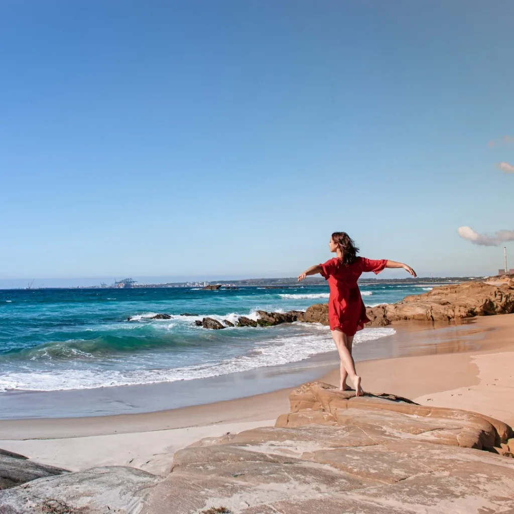 Caroline in a red dress walks on the rocks by the beach of Praia de Oliveirinha, near Porto Covo in Alentejo, under a clear sky in March 2022.